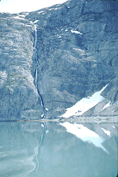 tall steep rocky cliff with long waterfall reflecting in calm waters in alaska