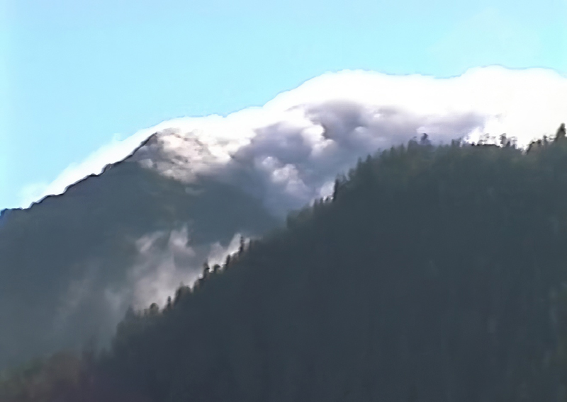 clouds creeping over top of mountain with trees outlined on dark hill in front