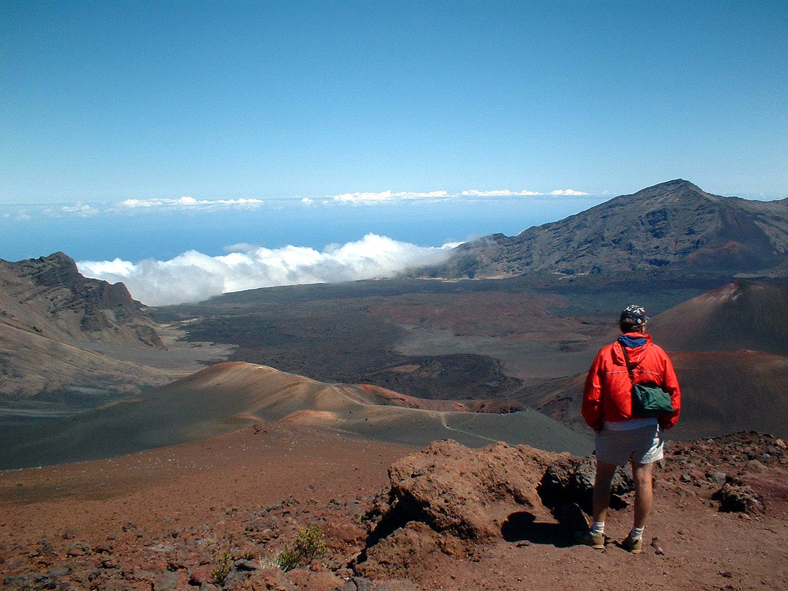 red orange brown sand and craters, blue sky, person in red jacket at top of haleakala national park maui hawaii