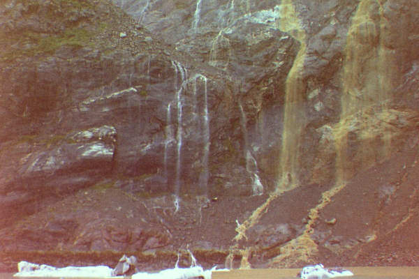 white waterfall and muddy brown waterfall from cliff into ice berg filled water